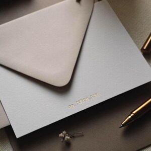 May include: A light grey rectangular card featuring the gold embossed text XO, NEOLANI, partially tucked into a taupe triangular envelope flap. The stationery set is arranged with a metallic gold pen and a small dried flower sprig nearby.
