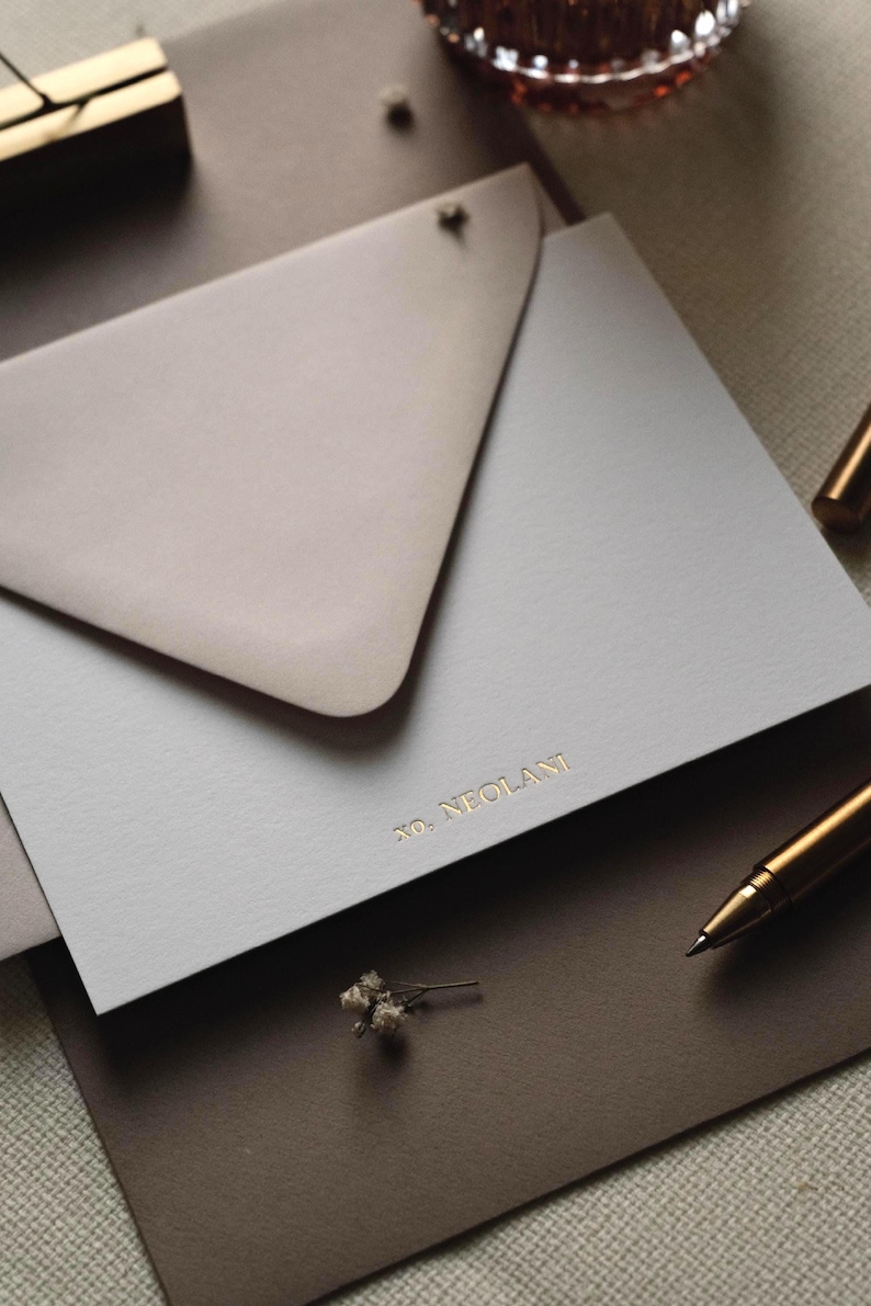 May include: A light grey rectangular card featuring the gold embossed text XO, NEOLANI, partially tucked into a taupe triangular envelope flap. The stationery set is arranged with a metallic gold pen and a small dried flower sprig nearby.