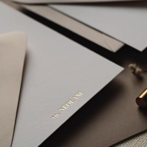 May include: A stack of stationery featuring light grey and tan envelopes and paper. The top light grey paper is embossed with the gold text "NO. NEOLANI" in a serif font. A small portion of a metallic gold pen is visible on the right side.