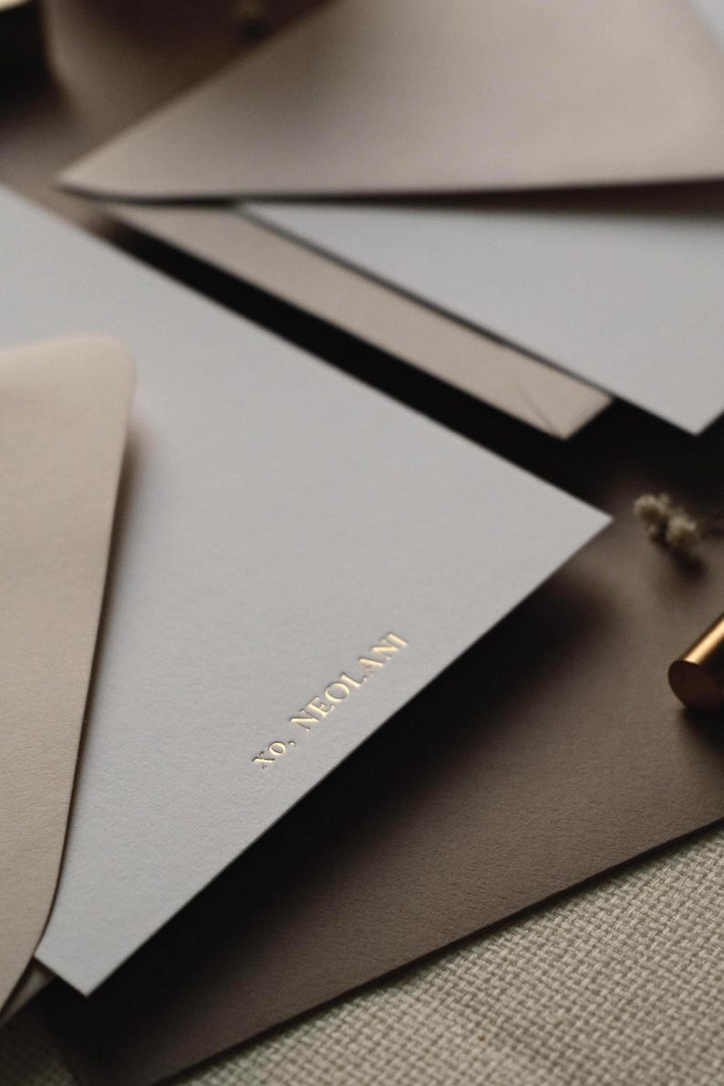 May include: A stack of stationery featuring light grey and tan envelopes and paper. The top light grey paper is embossed with the gold text "NO. NEOLANI" in a serif font. A small portion of a metallic gold pen is visible on the right side.