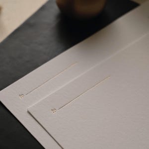 Puede incluir: Dos tarjetas rectangulares de color blanco roto con una superficie de papel texturizado. Cada tarjeta muestra la palabra to en una peque&ntilde;a fuente serif dorada, seguida de una fina l&iacute;nea horizontal dorada para escribir el nombre del destinatario.