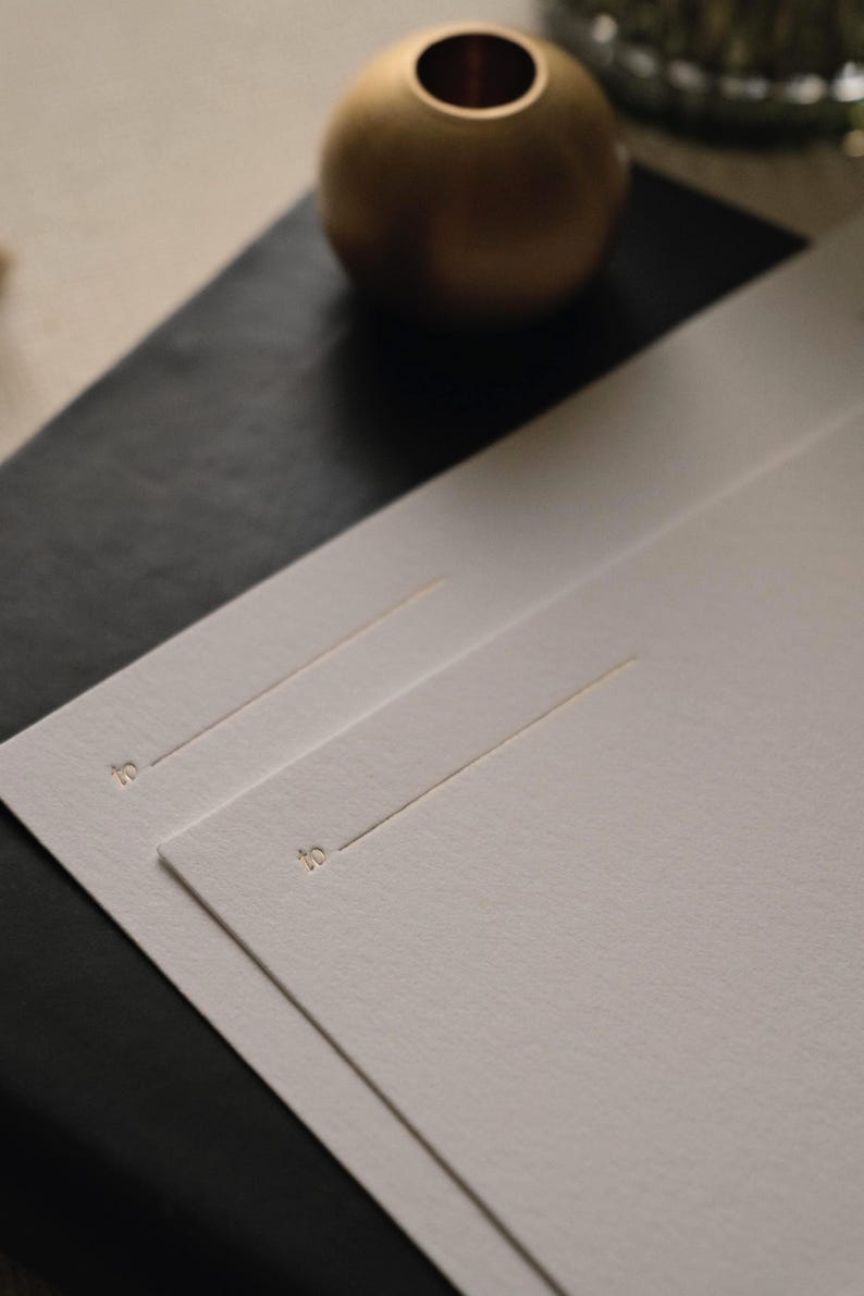 Puede incluir: Dos tarjetas rectangulares de color blanco roto con una superficie de papel texturizado. Cada tarjeta muestra la palabra to en una peque&ntilde;a fuente serif dorada, seguida de una fina l&iacute;nea horizontal dorada para escribir el nombre del destinatario.