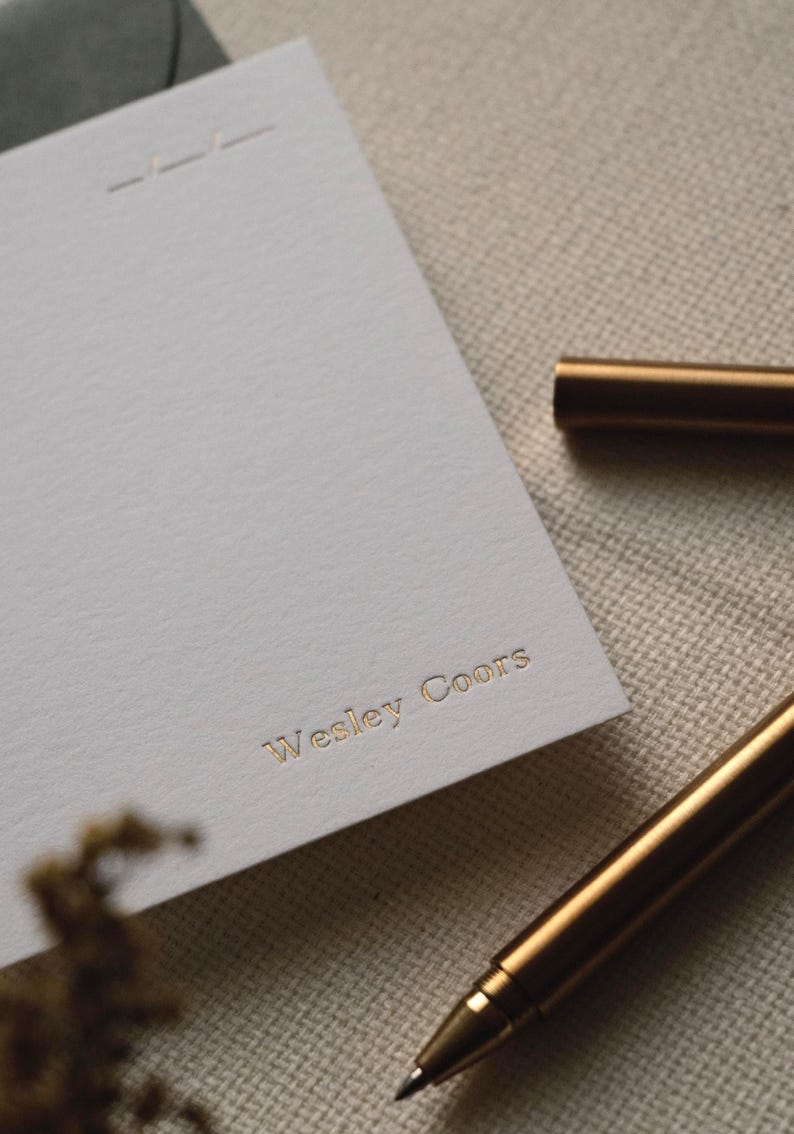 Puede incluir: Una tarjeta de papeler&iacute;a rectangular blanca con el nombre Wesley Coors grabado en letras de papel de oro. La tarjeta incluye un detalle de l&iacute;nea horizontal sutil en la parte superior y se coloca junto a un bol&iacute;grafo dorado met&aacute;lico sobre una tela.