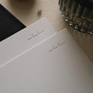Puede incluir: Dos hojas apiladas de papel blanco grueso y texturizado. Cada hoja presenta un peque&ntilde;o logotipo geom&eacute;trico grabado cerca de la esquina superior derecha, que consiste en tres l&iacute;neas horizontales cortas en un patr&oacute;n escalonado.