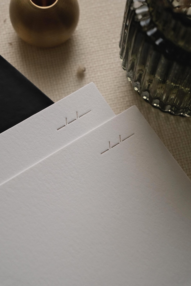 Puede incluir: Dos hojas apiladas de papel blanco grueso y texturizado. Cada hoja presenta un peque&ntilde;o logotipo geom&eacute;trico grabado cerca de la esquina superior derecha, que consiste en tres l&iacute;neas horizontales cortas en un patr&oacute;n escalonado.