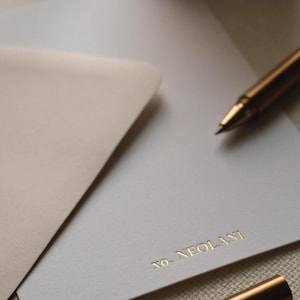 May include: A white stationery card featuring the gold embossed text XO, NEOLANI at the bottom right corner. The card is partially tucked into a light beige envelope, accompanied by a metallic gold pen resting on the paper surface.