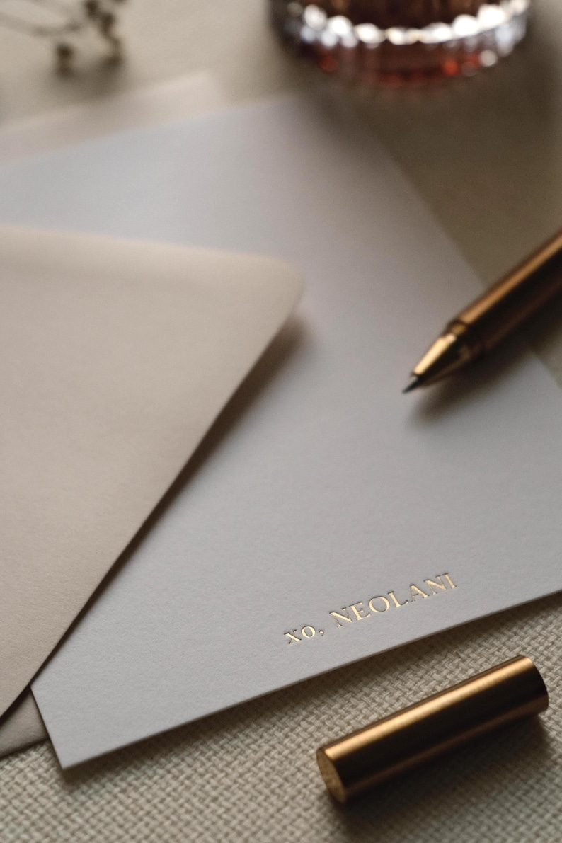 May include: A white stationery card featuring the gold embossed text XO, NEOLANI at the bottom right corner. The card is partially tucked into a light beige envelope, accompanied by a metallic gold pen resting on the paper surface.