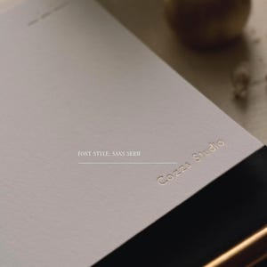 Puede incluir: Una tarjeta de papel rectangular de color gris claro con el texto dorado en relieve Cozzs Studio en una fuente sans serif. La tarjeta tiene una textura mate y lisa, y est&aacute; colocada en &aacute;ngulo, destacando las letras met&aacute;licas sobre la superficie.