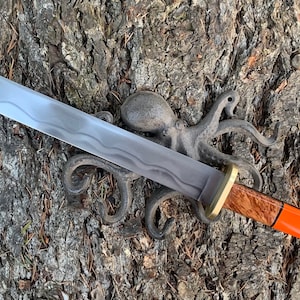 May include: A long, silver-colored blade with a wooden handle and a black and orange accent. The blade has a wavy pattern and is resting on a tree trunk with a metal octopus sculpture.