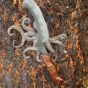 May include: A black and silver knife with a wooden handle. The handle has a yellow stripe. The knife is resting on a piece of wood with a decorative octopus design.