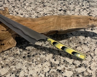 May include: A black and yellow patterned knife with a long, thin blade. The knife is resting on a piece of driftwood.