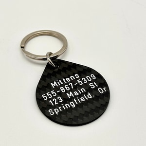 May include: Black carbon fiber keychain with a teardrop-shaped tag. The tag is engraved with the text "Mittens 555-867-5309 123 Main St Springfield. Or"