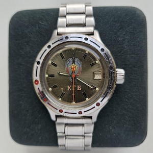 Puede incluir: Un reloj de pulsera plateado con una esfera redonda que muestra un emblema de escudo y espada, las letras "KGB" y una ventana de fecha. El reloj tiene una correa plateada y un bisel giratorio con detalles en rojo. El reloj se muestra sobre una superficie verde oscuro.