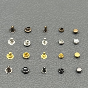 May include: A collection of various metal rivets in different sizes and finishes, including gold, silver, bronze, and black. The rivets are arranged on a gray surface, showcasing their diverse shapes and styles.