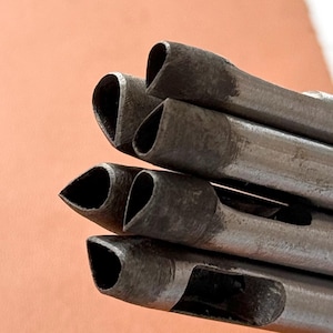 Könnte beinhalten: Nahaufnahme eines Satzes von Metall-Lederwerkzeugen. Die Werkzeuge haben eine dunkle, metallische Oberfläche und eine einzigartige, abgewinkelte Schneidkante. Die Werkzeuge werden vor einer hellbraunen Lederoberfläche gehalten.