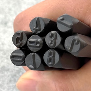 May include: A collection of dark metal number stamps, featuring the digits 0 through 9. The stamps are held together, with the numbers clearly visible. The tools are likely used for marking or imprinting numbers on various materials.