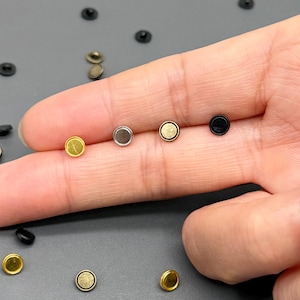 May include: A selection of small, circular metal rivets in assorted colours, including gold, silver, and black. The rivets are scattered on a grey surface, with some held in a hand. These fasteners are likely used for crafting or leatherwork.