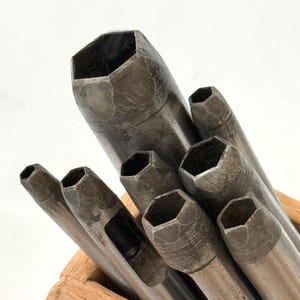 Op de afbeelding: Een verzameling metalen gatenponsen met zeshoekige koppen, in verschillende maten, is gerangschikt in een houten kist. De ponsen hebben een versleten, industriële uitstraling, wat duidt op veelvuldig gebruik. De gereedschappen zijn schuin geplaatst, waardoor een dynamische compositie ontstaat tegen een witte achtergrond.