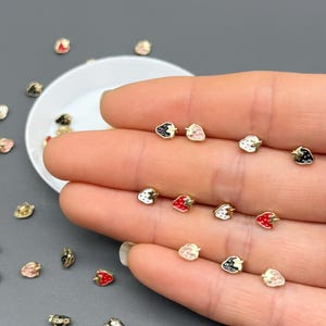 May include: Tiny strawberry-shaped stud earrings in various colors, including red, black, white, and pink. The earrings have a gold-colored metal base. Some earrings are scattered on a gray surface, while others are held in a hand.