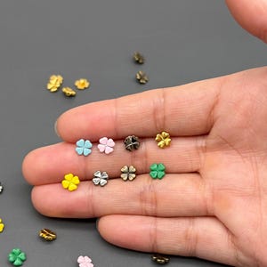May include: A collection of small, four-leaf clover charms in various colors, including gold, silver, black, yellow, pink, blue, and green. The charms are scattered on a gray surface, with some held in a hand.