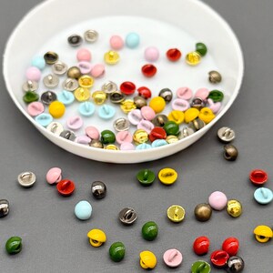 Puede incluir: Una colección de pequeños botones coloridos en varios tonos, incluyendo rosa, amarillo, verde, rojo y azul. Algunos botones son metálicos, mientras que otros son de colores sólidos. Los botones están dispersos sobre una superficie gris y en un cuenco blanco.