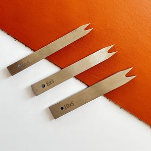May include: Three metal leather working tools with a forked end, arranged diagonally on an orange leather surface. Each tool is marked with a size: 6x4, 8x4, and 10x5. The background is a bright orange leather.
