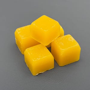 Può includere: Primo piano di cinque cubetti di cera gialla a forma di cubo. I cubetti di cera sono impilati, con un aspetto leggermente traslucido. I cubetti hanno una superficie liscia e sono di colore giallo brillante.