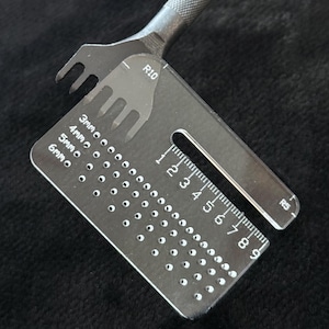 May include: A metal leathercraft tool set, featuring a pronged punch and a clear acrylic guide with measurements in millimeters and a ruler. The tool is designed for precision leatherwork, with markings for various hole sizes.