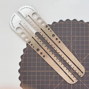 May include: Two silver metal angle finders with measurement markings and various hole sizes. The tools are positioned diagonally on a dark grey cutting mat with a scalloped edge. The tools have a series of holes and markings for precise measurements.