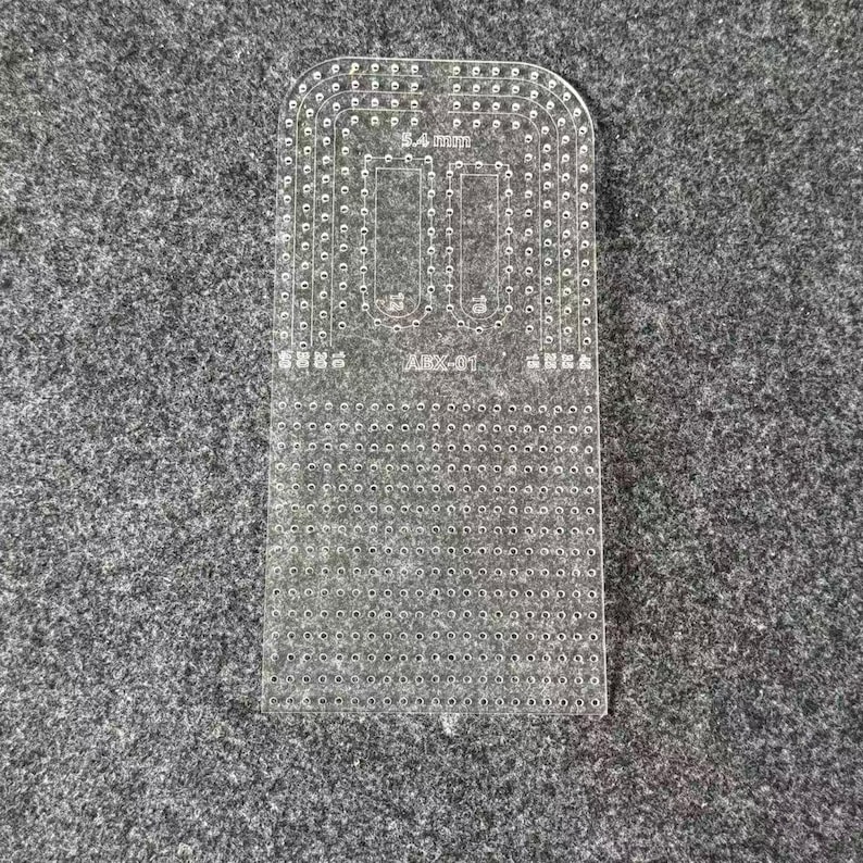 Peut inclure: Un gabarit de maroquinerie en acrylique transparent dot&eacute; d'une grille de petits trous pour la couture, avec des coins sup&eacute;rieurs arrondis. L'outil porte l'inscription 5.4 mm et ABX-01, ainsi que divers indicateurs num&eacute;riques pour l'espacement.
