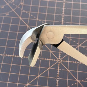 Puede incluir: Primer plano de una herramienta de metal plateado con mordazas curvas y un mango articulado. La herramienta parece ser un sacaclavos o un tipo similar de herramienta de palanca. El fondo es una superficie oscura con un patr&oacute;n de cuadr&iacute;cula.