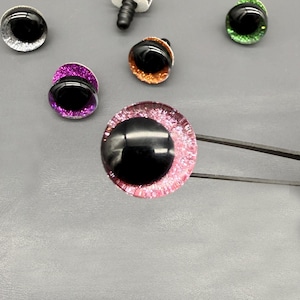 Puede incluir: Una colección de ojos artesanales coloridos y brillantes con pupilas negras. Los ojos varían en color, incluyendo plata, púrpura, naranja, verde y rosa. Un ojo es sostenido por unas pinzas. Ideales para manualidades y fabricación de muñecas.