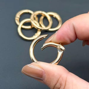 Könnte beinhalten: Eine Sammlung von goldfarbenen Metallringen mit Federverschluss. Jeder kreisförmige Ring verfügt über ein aufklappbares Segment, das sich öffnen und schließen lässt, um Anhänger oder andere Gegenstände zu befestigen. Das Metall ist glatt poliert.