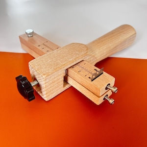 May include: A wooden leather edge creaser tool with a blade and adjustable settings. The tool has a wooden handle and a black knob for adjustments. The tool is designed for leathercraft and has measurement markings in inches.