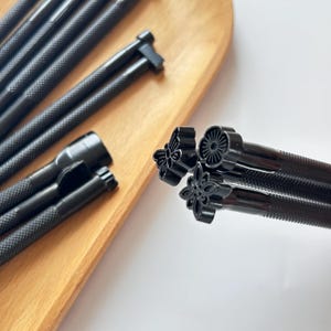 Pode incluir: Close-up de várias ferramentas de estampagem de couro em metal preto. As ferramentas têm cabos texturizados e vários desenhos florais e geométricos nas cabeças de estampagem. Algumas ferramentas estão em uma tábua de madeira, outras em uma superfície branca.