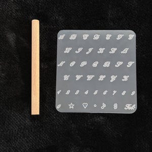 May include: A light brown wooden dowel next to a clear, rectangular stamp with white cursive letters, numbers, and symbols. The stamp includes the letters A through Z, numbers 1 through 9, and several symbols.