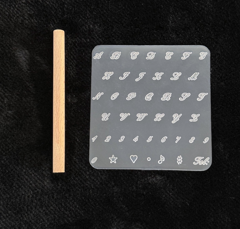 May include: A light brown wooden dowel next to a clear, rectangular stamp with white cursive letters, numbers, and symbols. The stamp includes the letters A through Z, numbers 1 through 9, and several symbols.