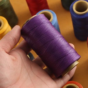 May include: A close-up of a hand holding a spool of purple thread. Other spools of thread in various colors, including red, blue, green, and orange, are visible in the background. The thread appears to be tightly wound and ready for use.