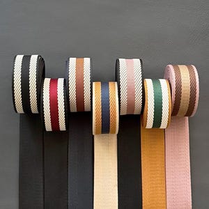 Puede incluir: Una colección de rollos de cinta a rayas en varios colores, incluyendo negro, burdeos, marrón, dorado, rosa y verde. Cada rollo presenta una combinación de colores diferente con rayas verticales y una textura tejida. Las cintas están desenrolladas en la parte inferior.