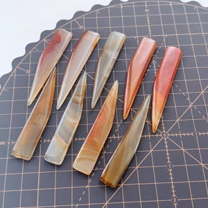 Puede incluir: Una colección de nueve herramientas de piedra pulida y alargada. Las herramientas son de colores cálidos, incluyendo tonos de marrón, naranja y crema, con patrones de vetas naturales. Están dispuestas sobre una superficie oscura.