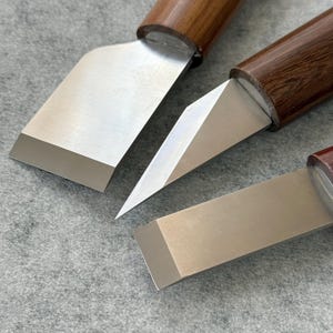 Peut inclure: Gros plan sur trois ciseaux à bois. Chaque ciseau est doté d'une lame en acier poli et d'un manche en bois brun foncé. Les lames ont des formes différentes, notamment un bord plat et un bord biseauté, ce qui met en valeur la polyvalence des outils.