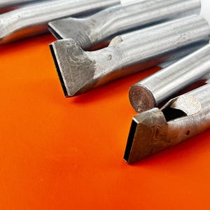May include: Close-up of several metal leather working tools with angled, rectangular cutting edges. The tools are arranged on an orange surface. The tools have a cylindrical handle and a flat, beveled blade. The tools are likely used for cutting and shaping leather.
