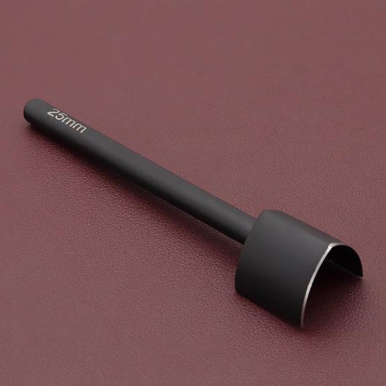 May include: A black leathercraft tool with a curved cutting edge. The tool has a long, cylindrical handle with the measurement "25mm" printed on it. The tool is designed for cutting leather and other materials. The tool is shown against a maroon background.