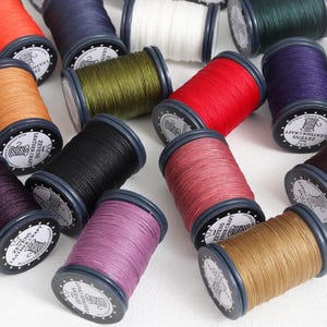 May include: A collection of colorful spools of thread, including red, black, and purple. Each spool is labeled with "ZEVENS LEATHER CRAFT" and features a dark gray base. The threads are tightly wound and appear ready for use in various crafting projects.