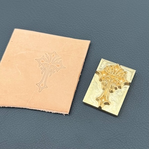 May include: A rectangular brass metal stamp featuring an intricate embossed cross design, positioned next to a square piece of light tan leather that displays a clear, pressed impression of the same cross pattern in its center.