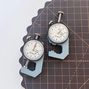 May include: Two light blue thickness gauges with white dials and black markings. The dials read "TAI NUO" and "0.1mm". The gauges are on a dark brown cutting mat with grid lines.