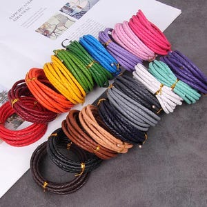 May include: A collection of braided leather bracelets in various colors, including red, orange, yellow, green, blue, purple, pink, white, gray, brown, and black. Each bracelet is coiled and tied with gold-colored string.