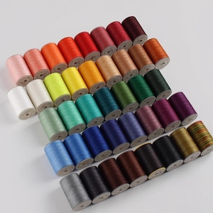 May include: A collection of colorful spools of thread, arranged in neat rows. The threads are in a variety of colors, including pink, orange, red, yellow, green, blue, brown, and purple. The spools are made of a smooth material and are tightly wound.