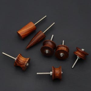 Peut inclure: Une collection de boutons et poignées de tiroir en bois faits à la main de différentes formes. Les boutons et poignées sont en bois brun foncé avec un motif de grain visible et comportent des vis en métal argenté. Les formes comprennent des designs cylindriques, coniques et en forme de bobine.
