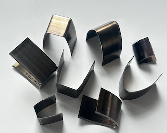 May include: A selection of metal leathercraft tools, including U-shaped and curved cutting dies. The tools are made of dark metal and are arranged on a white surface. Some tools have stamped markings.